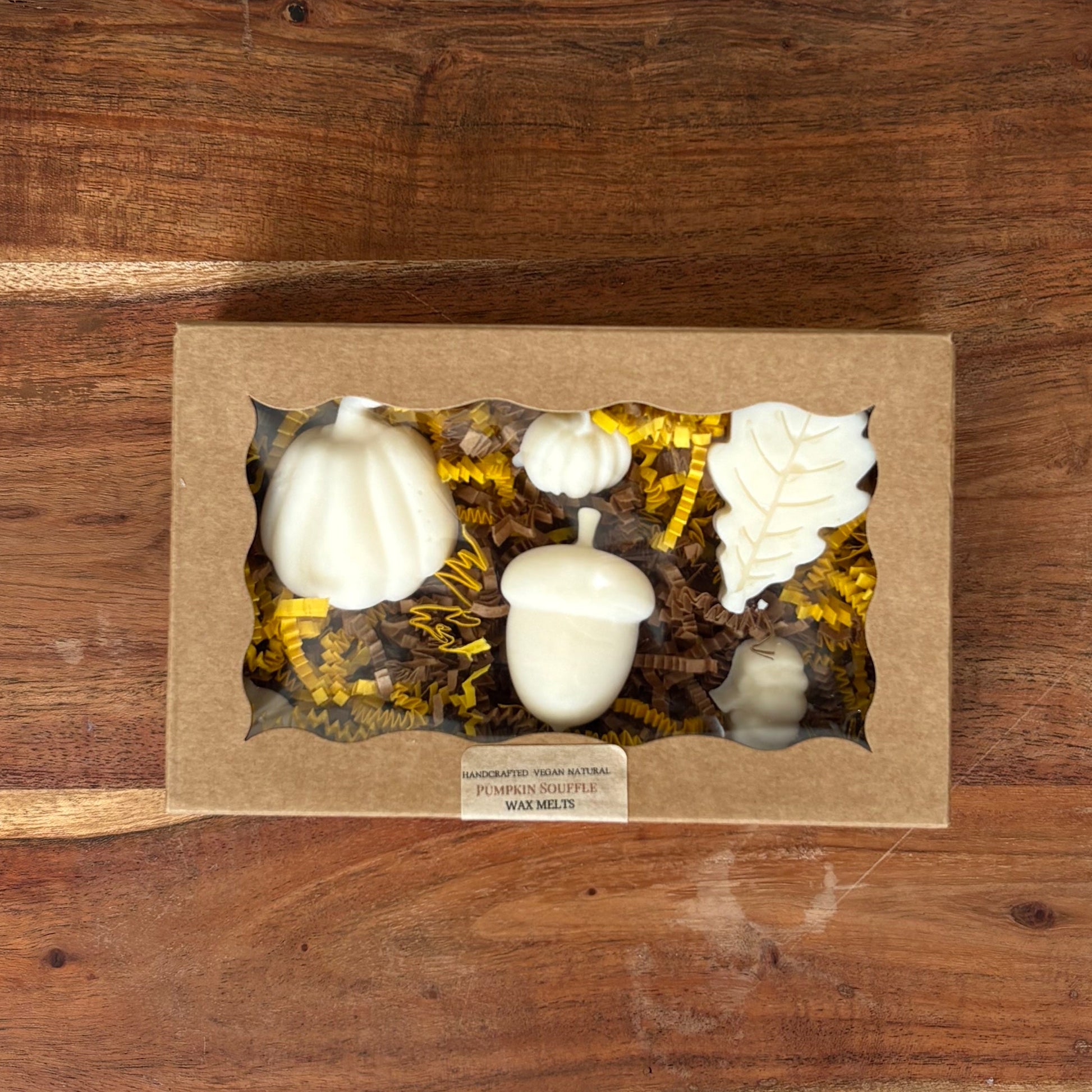 A decorative fall-themed gift box placed on a large wooden cutting board. The box contains wax melt pumpkins, acorns, and other autumn decorations nestled in yellow and brown faux foliage.