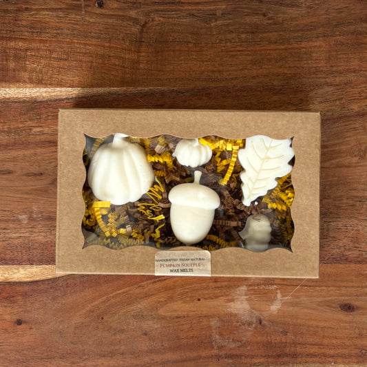 A decorative fall-themed gift box placed on a large wooden cutting board. The box contains wax melt pumpkins, acorns, and other autumn decorations nestled in yellow and brown faux foliage.
